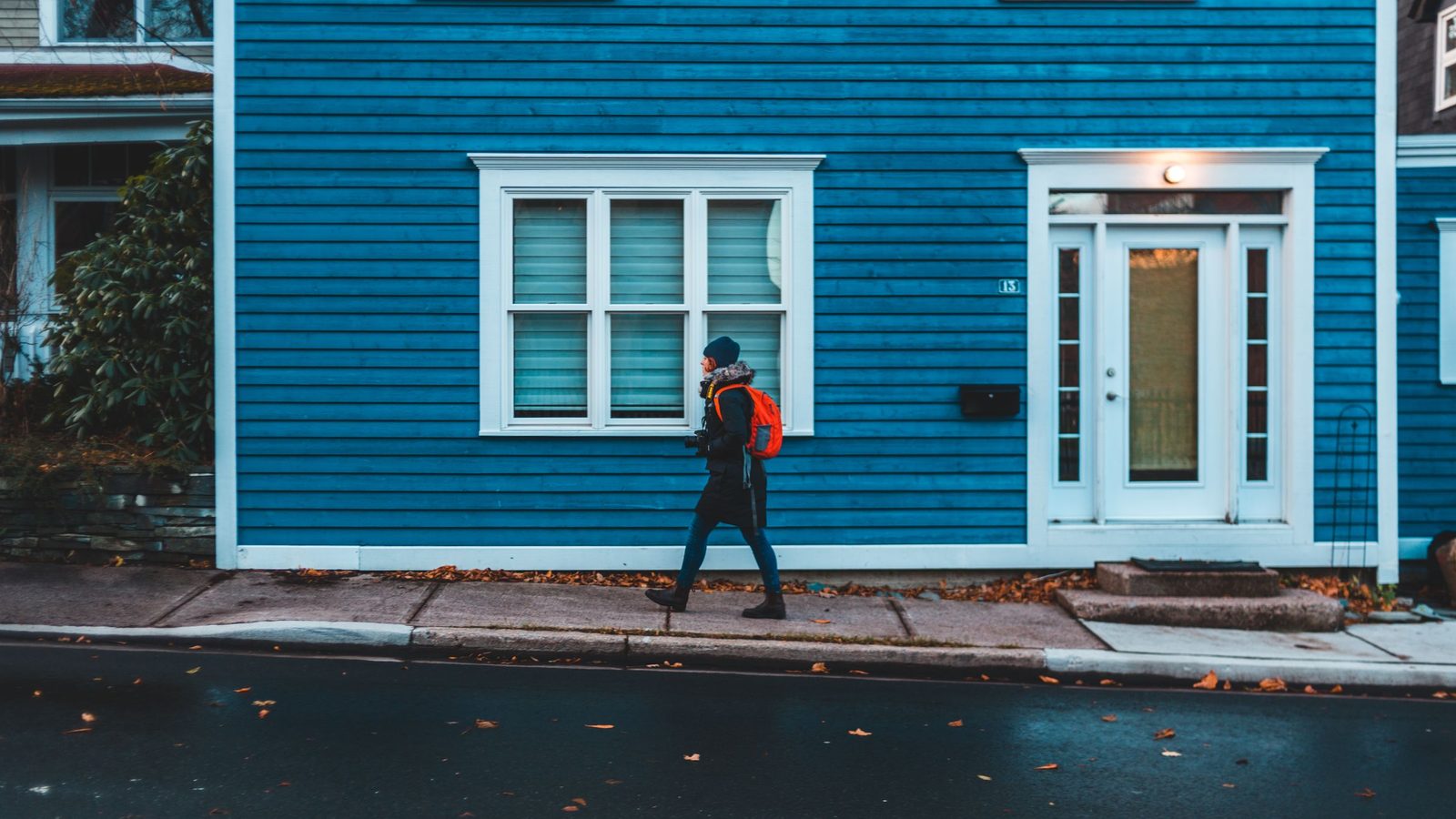 A person with a backpack walks along the sidewalk past a blue house