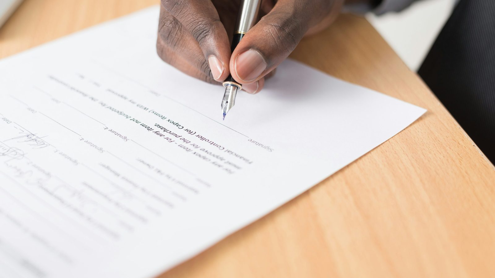 A hand signs a printed paper form with a pen on a wooden desk