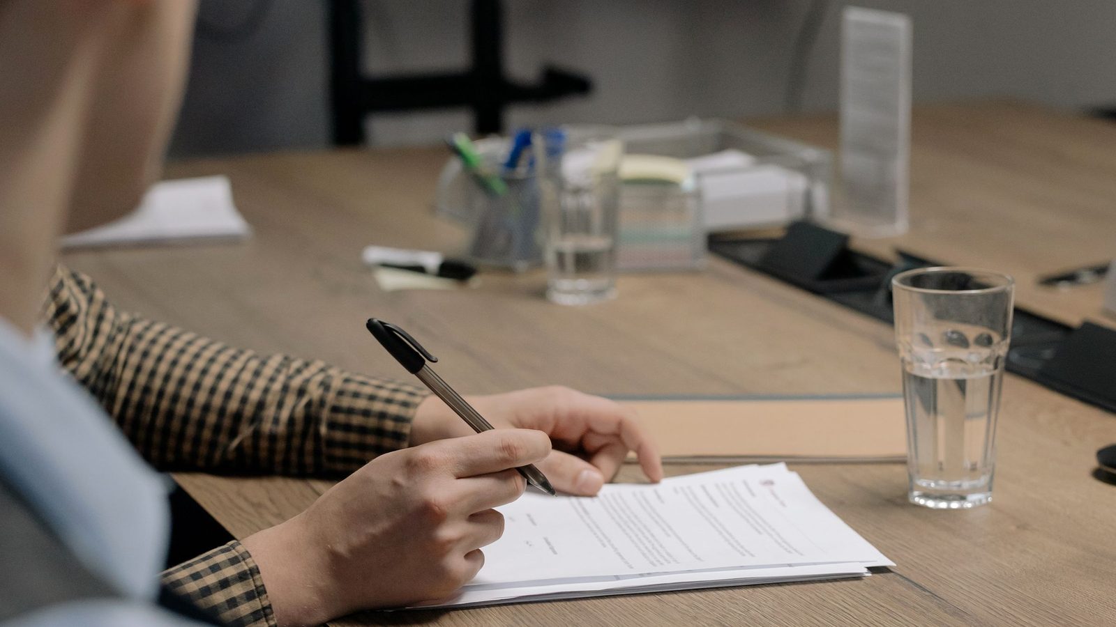 A person in a checked shirt signs a printed document at a wooden table