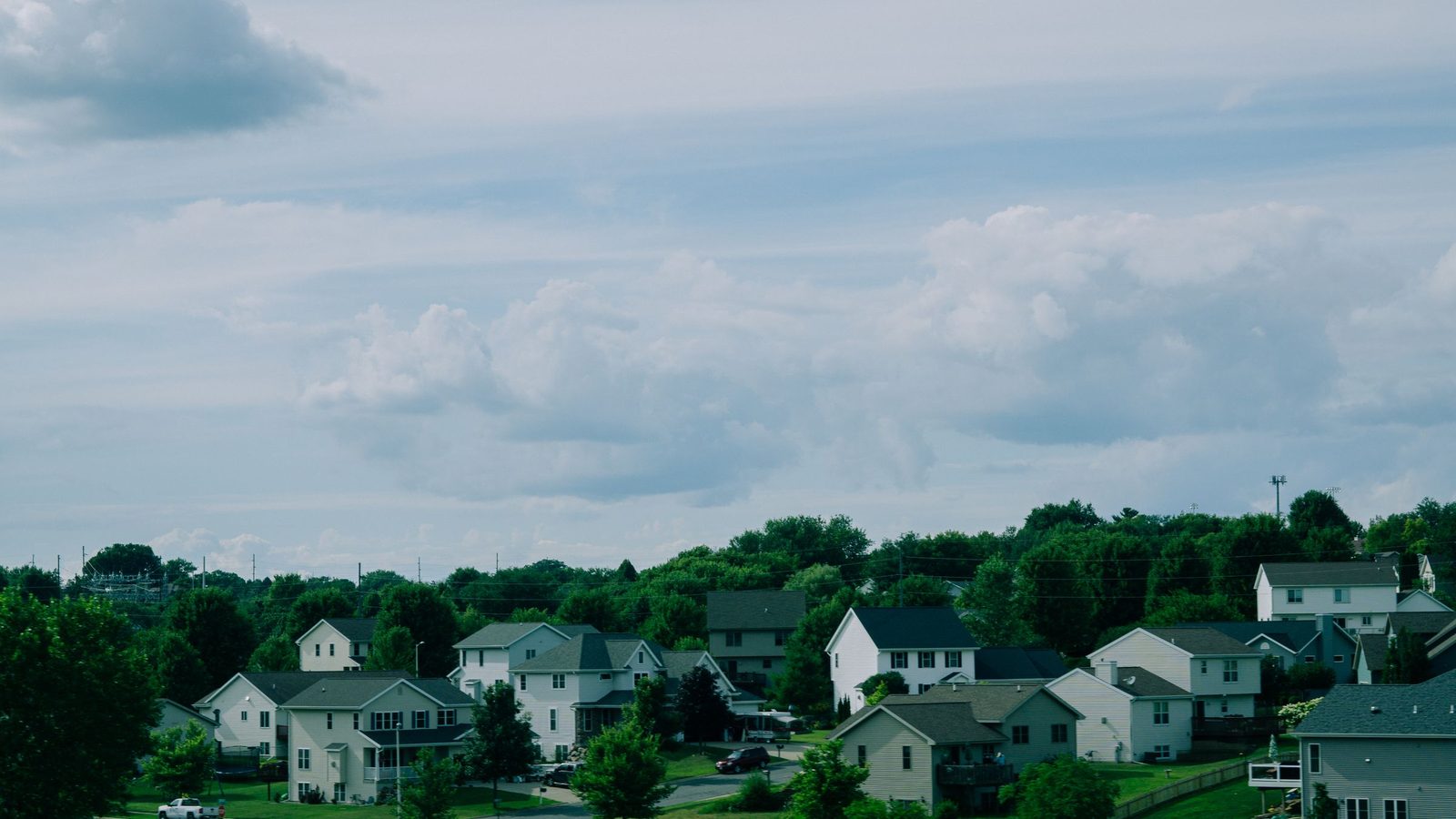 Rows of suburban houses among green trees under a cloudy blue sky