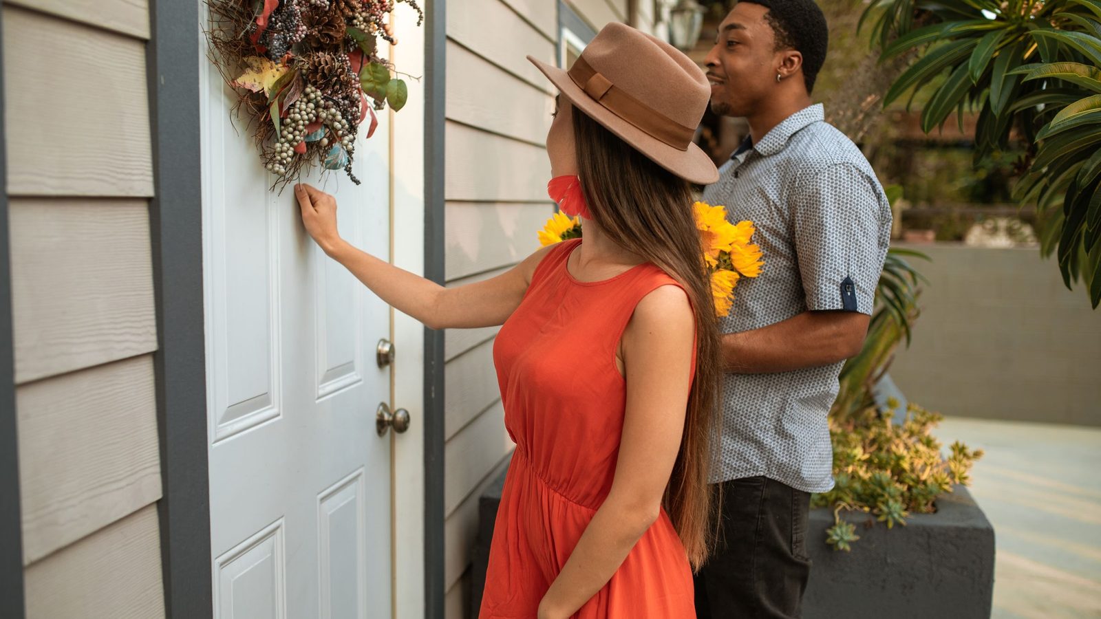 A man and a woman knock on a white front door decorated with a wreath