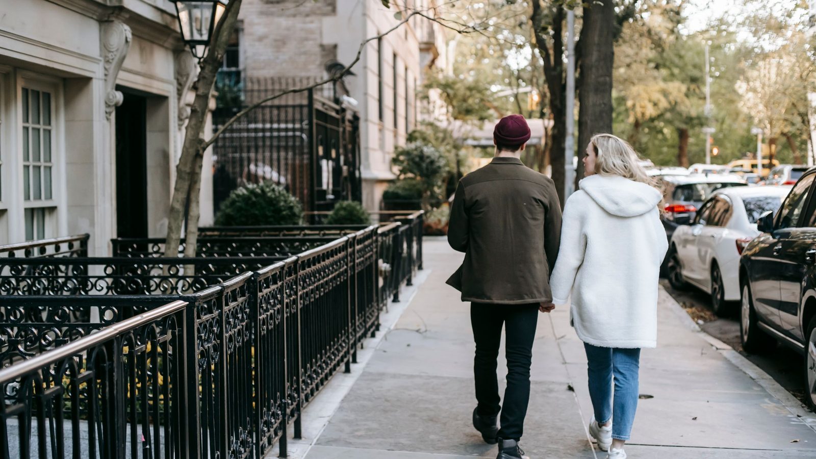 Two people walk side by side down a sidewalk lined with iron fences and parked cars