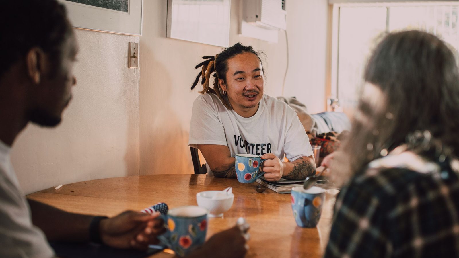 A man in a volunteer T-shirt talks with two people over coffee at a round table