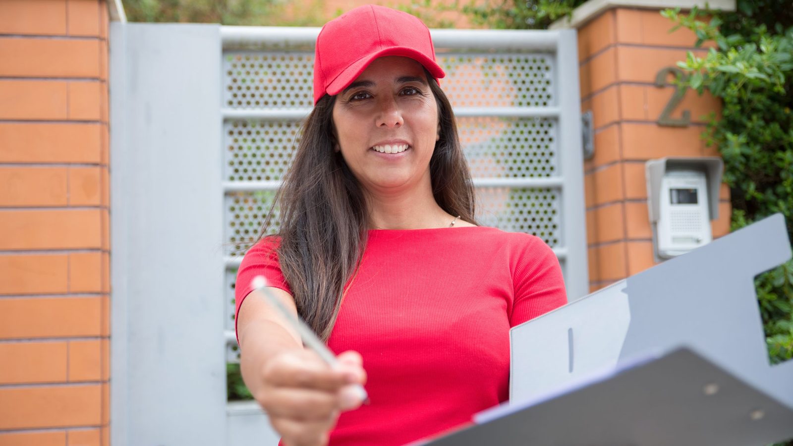 A smiling woman in a red cap holds out a pen and a clipboard at a front gate