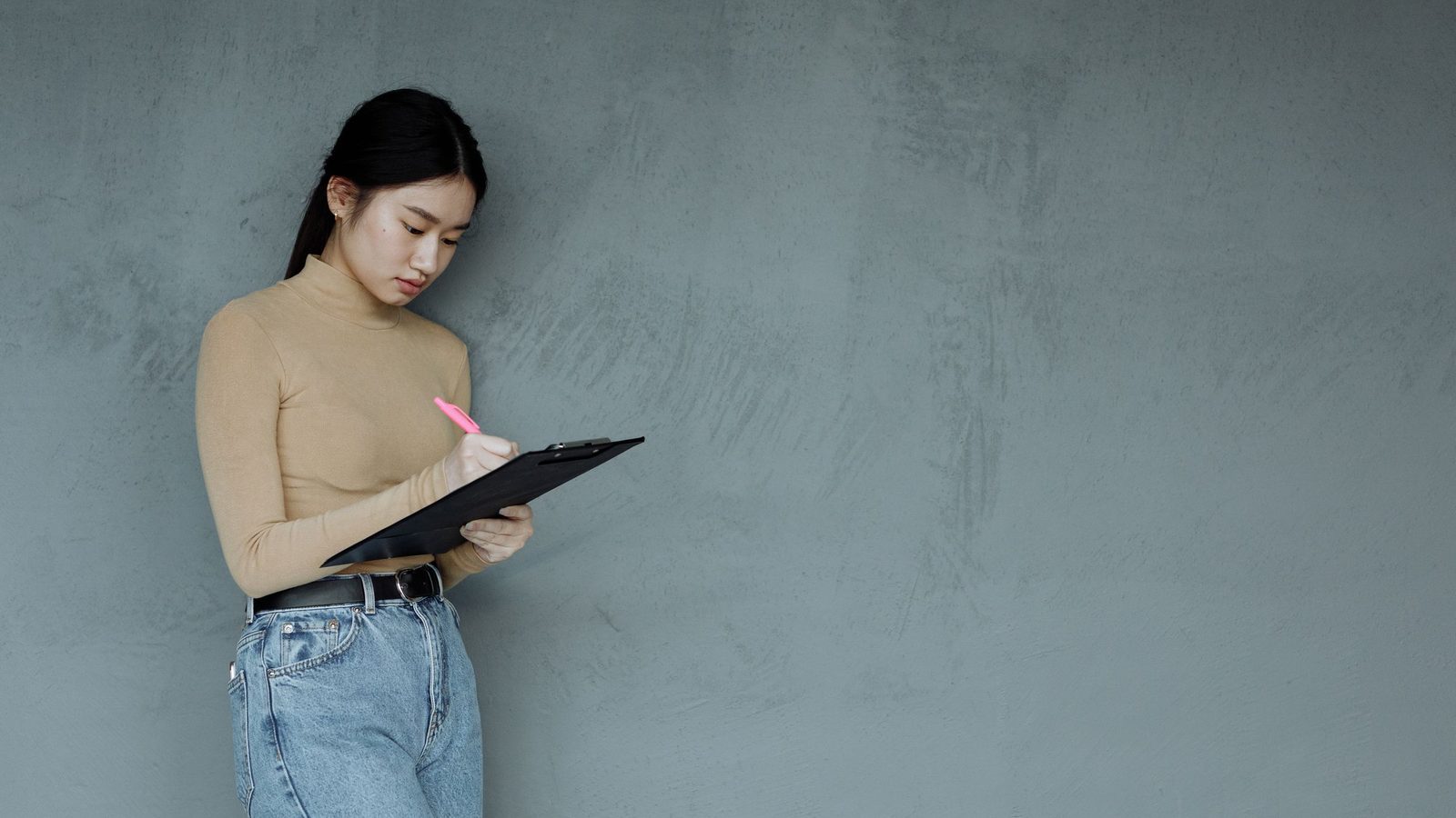 A woman writes on a clipboard while standing against a gray wall